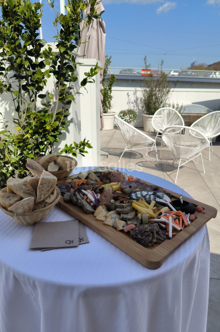 Plateau de charcuterie et fromages avec légumes et pain, posé sur une table dressée en extérieur, près de chaises et plantes.