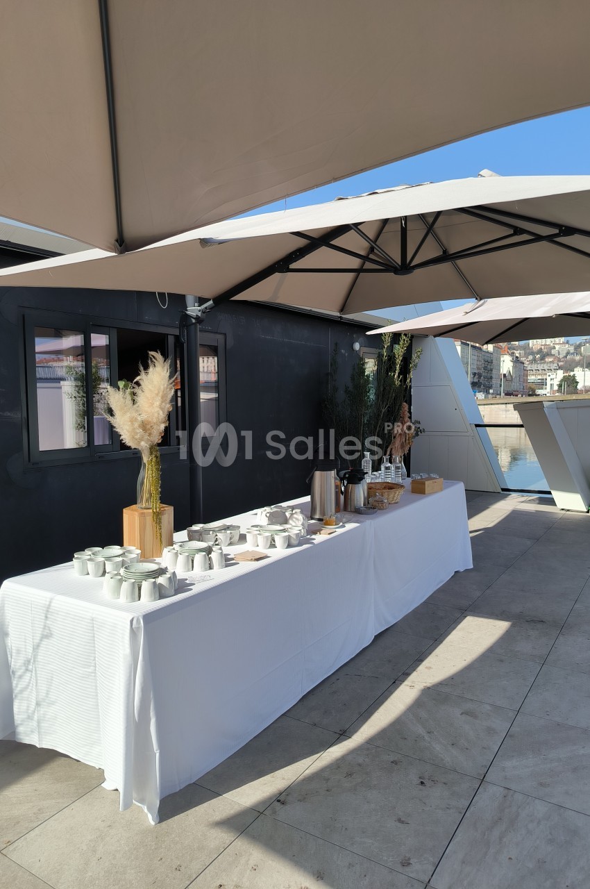 Table dressée en extérieur sous des parasols, avec vaisselle, thermos et décorations végétales.