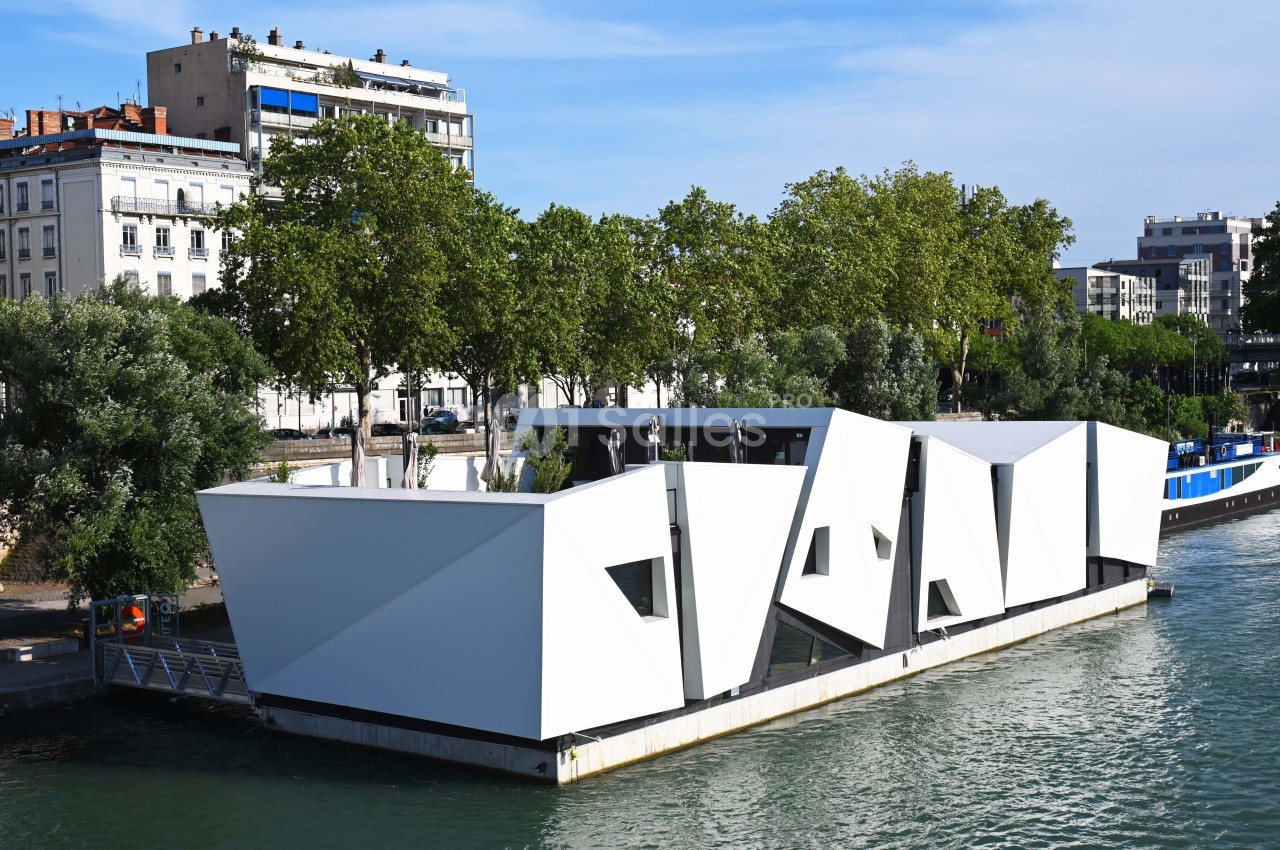 Bâtiment flottant moderne en structure blanche géométrique, amarré sur une rivière bordée d'arbres et d'immeubles.