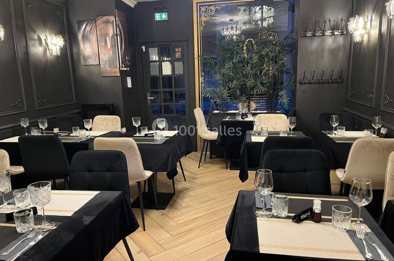 Salle de restaurant élégante avec tables dressées, chaises modernes et décoration sombre rehaussée de plantes et luminaires.
