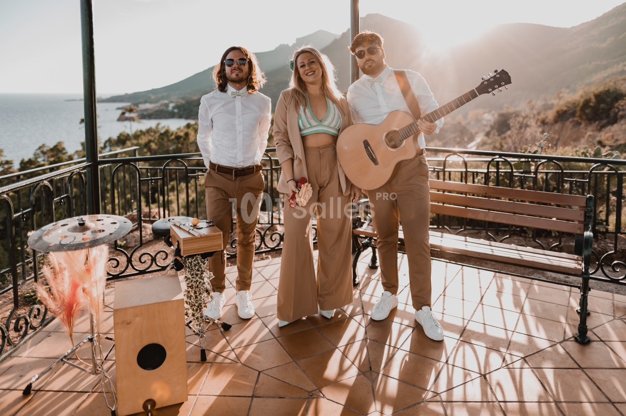 Trois musiciens posent sur une terrasse ensoleillée avec vue sur la mer et les montagnes, guitare et percussions visibles.