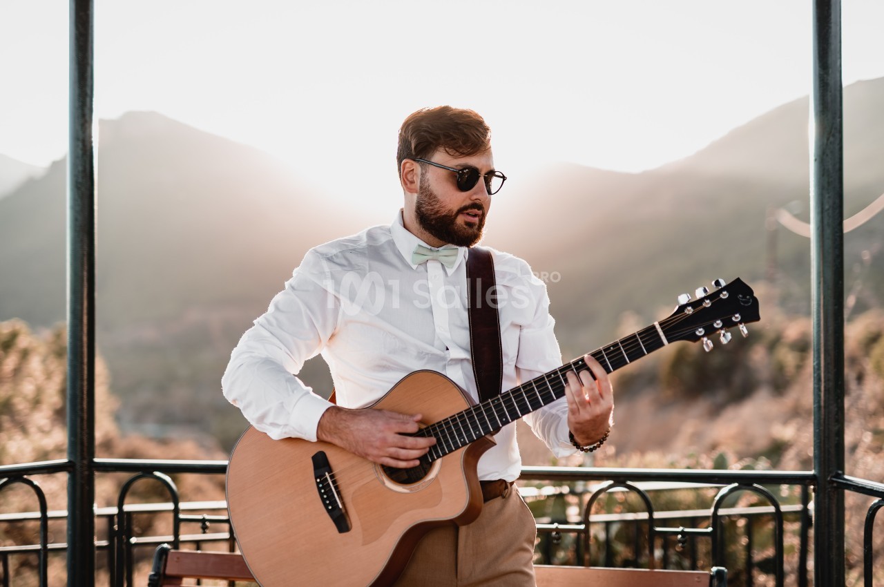 Un homme joue de la guitare acoustique sous un kiosque en plein air, avec des montagnes en arrière-plan.