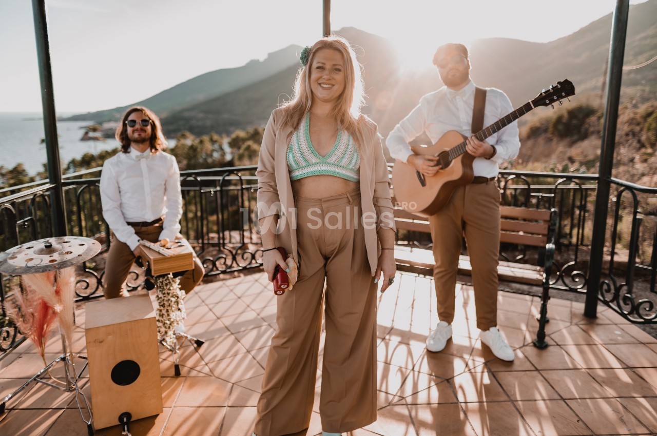 Une chanteuse souriante entourée de deux musiciens jouant de la guitare et du cajón sur une terrasse ensoleillée.