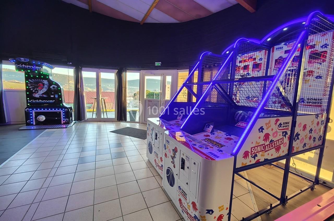 Jeu d'arcade de basketball lumineux dans une salle avec un autre jeu rétro en arrière-plan et des baies vitrées.