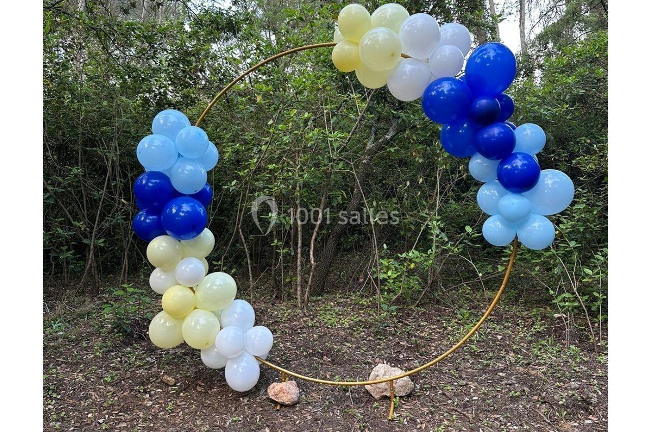 Cercle doré décoré de ballons bleus, blancs et jaunes, placé dans un environnement boisé.