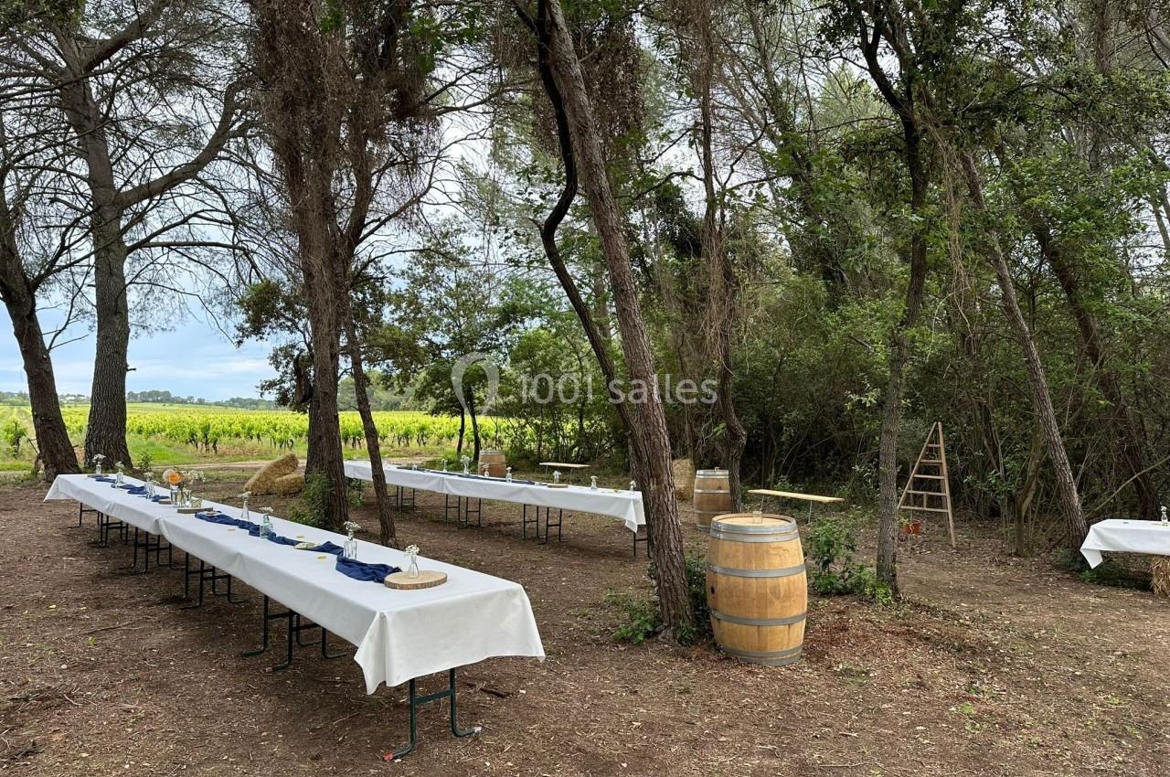 Tables dressées avec nappes blanches dans une clairière boisée, entourées de végétation et d'un tonneau en bois.