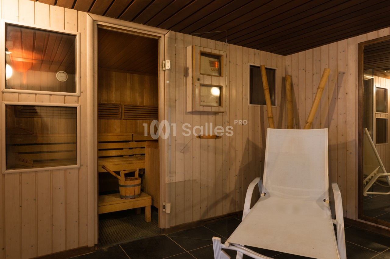 Sauna en bois avec bancs à l'intérieur, porte ouverte, chaise longue blanche et seau en bois au premier plan.
