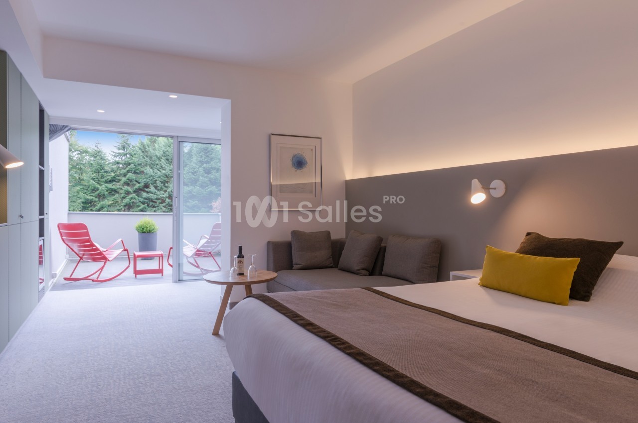 Chambre moderne avec lit double, canapé, table basse et balcon aménagé donnant sur des arbres.