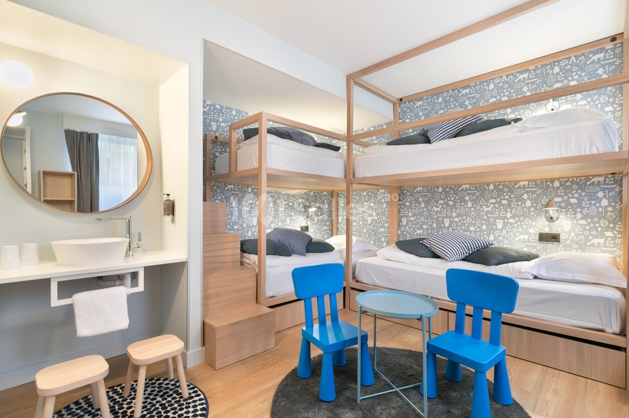 Chambre avec lits superposés en bois, coin lavabo, deux chaises bleues et une table ronde dans un décor moderne.