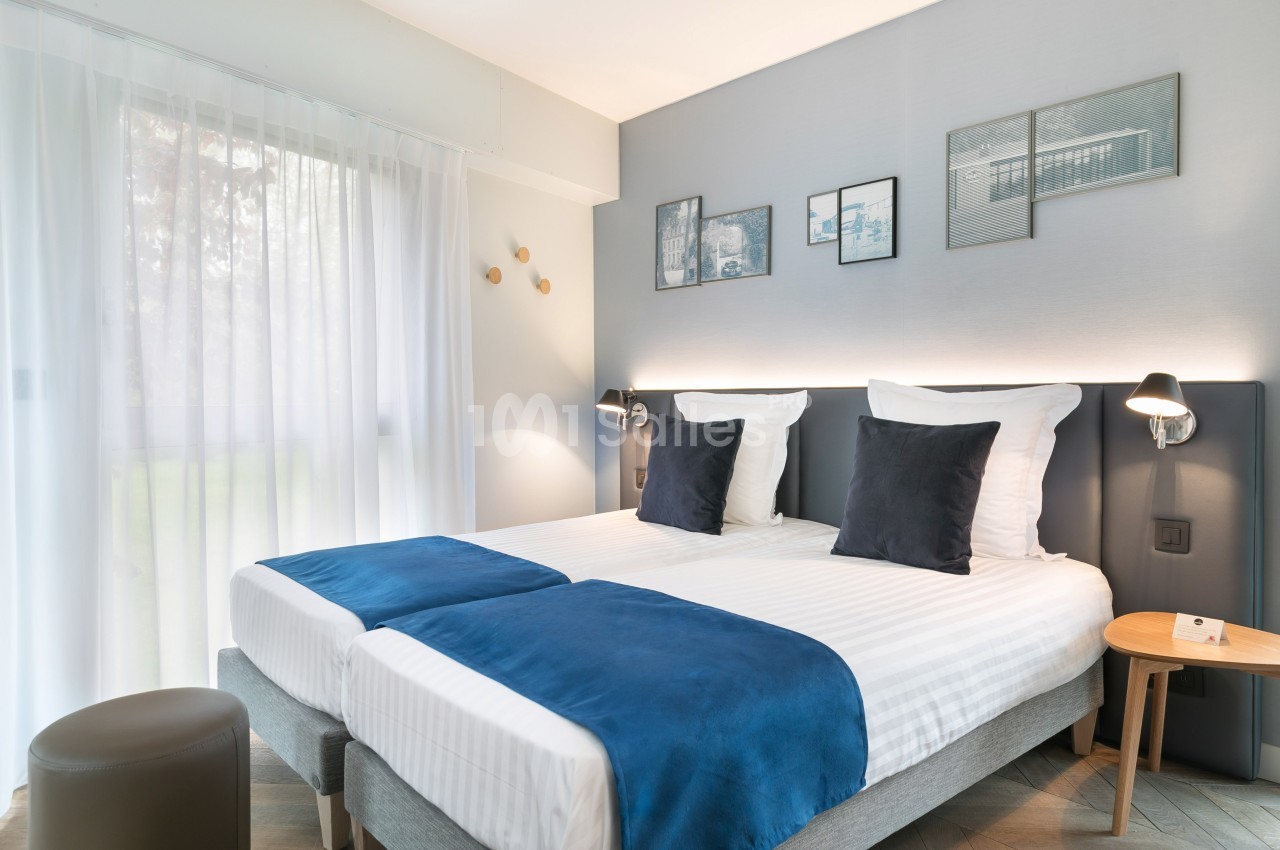 Chambre lumineuse avec deux lits simples, draps blancs, couvre-lits bleus, décor minimaliste et grande fenêtre avec rideaux.