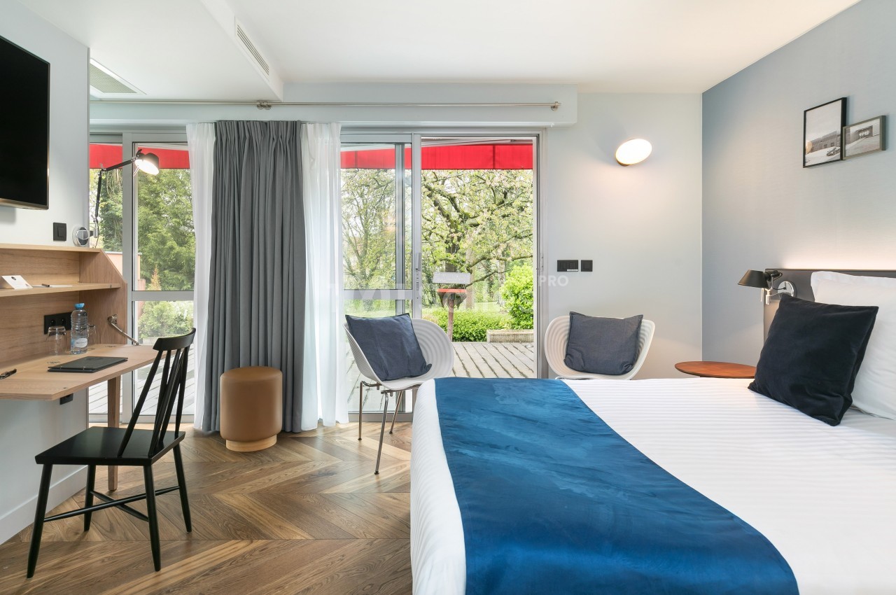 Chambre lumineuse avec lit double, bureau, fauteuils et grande baie vitrée donnant sur un balcon avec vue sur la verdure.