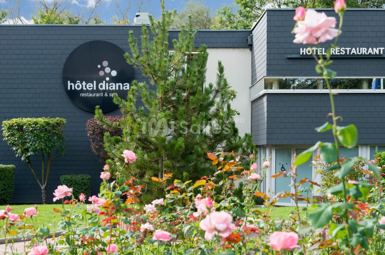 Façade d'un hôtel moderne entourée de rosiers en fleurs et d'arbres, avec un panneau indiquant ’hôtel diana’.