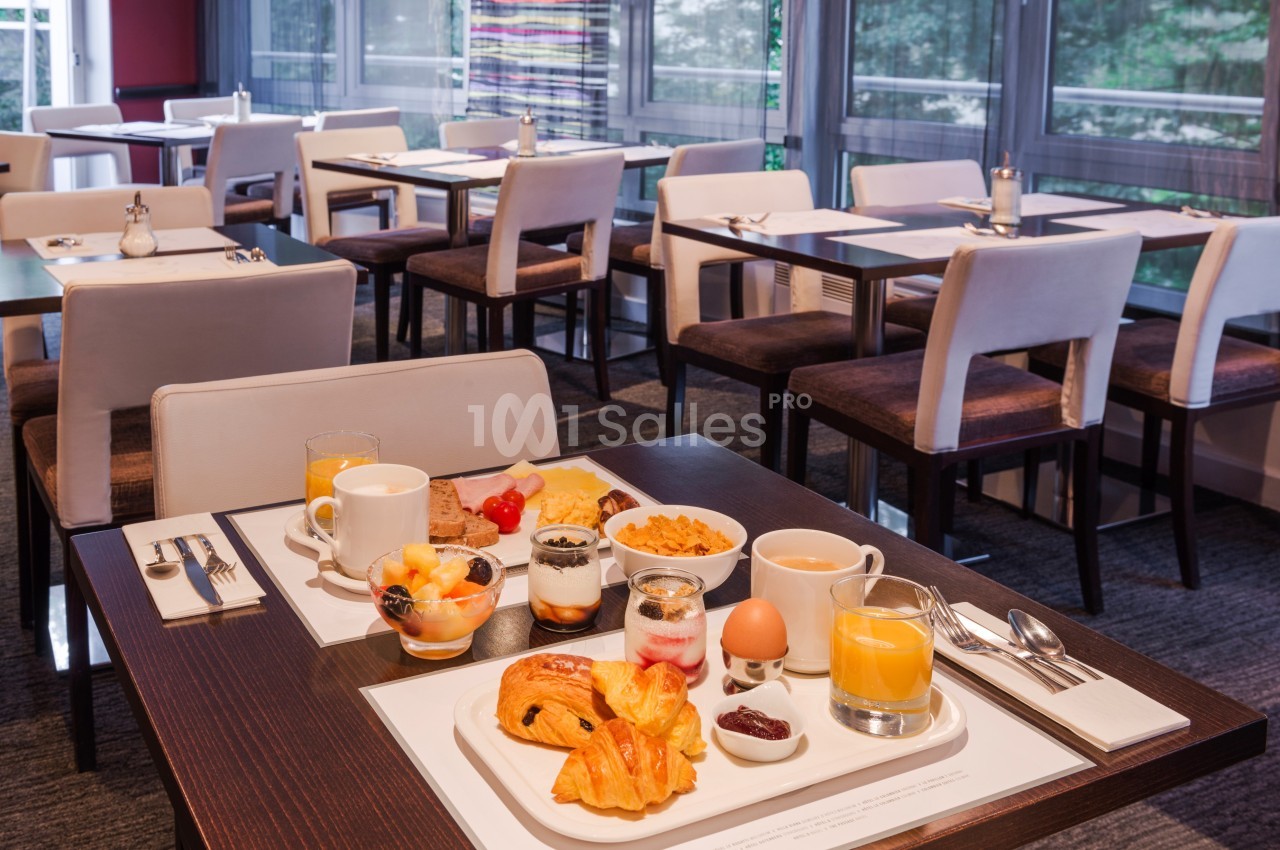 Petit-déjeuner servi sur une table avec viennoiseries, fruits, céréales, jus d'orange et yaourts, dans une salle lumineuse.