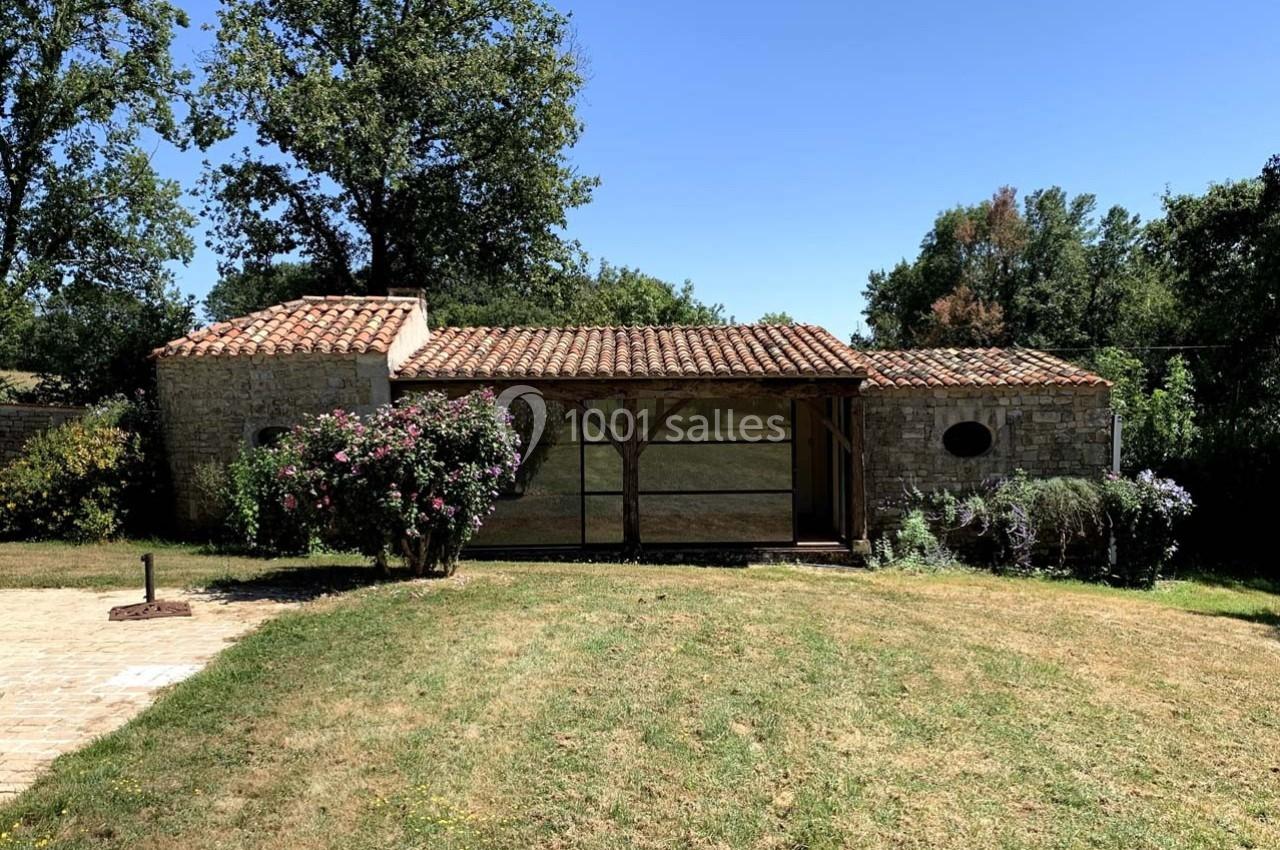 Maison en pierre avec toit en tuiles, entourée de végétation et d'un jardin arboré sous un ciel dégagé.
