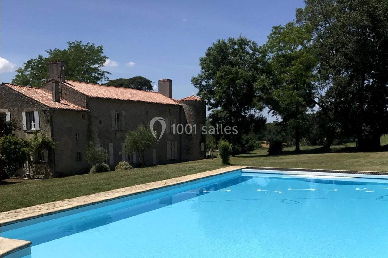 Grande piscine extérieure devant une maison en pierre avec toit en tuiles, entourée d'arbres et d'herbe.