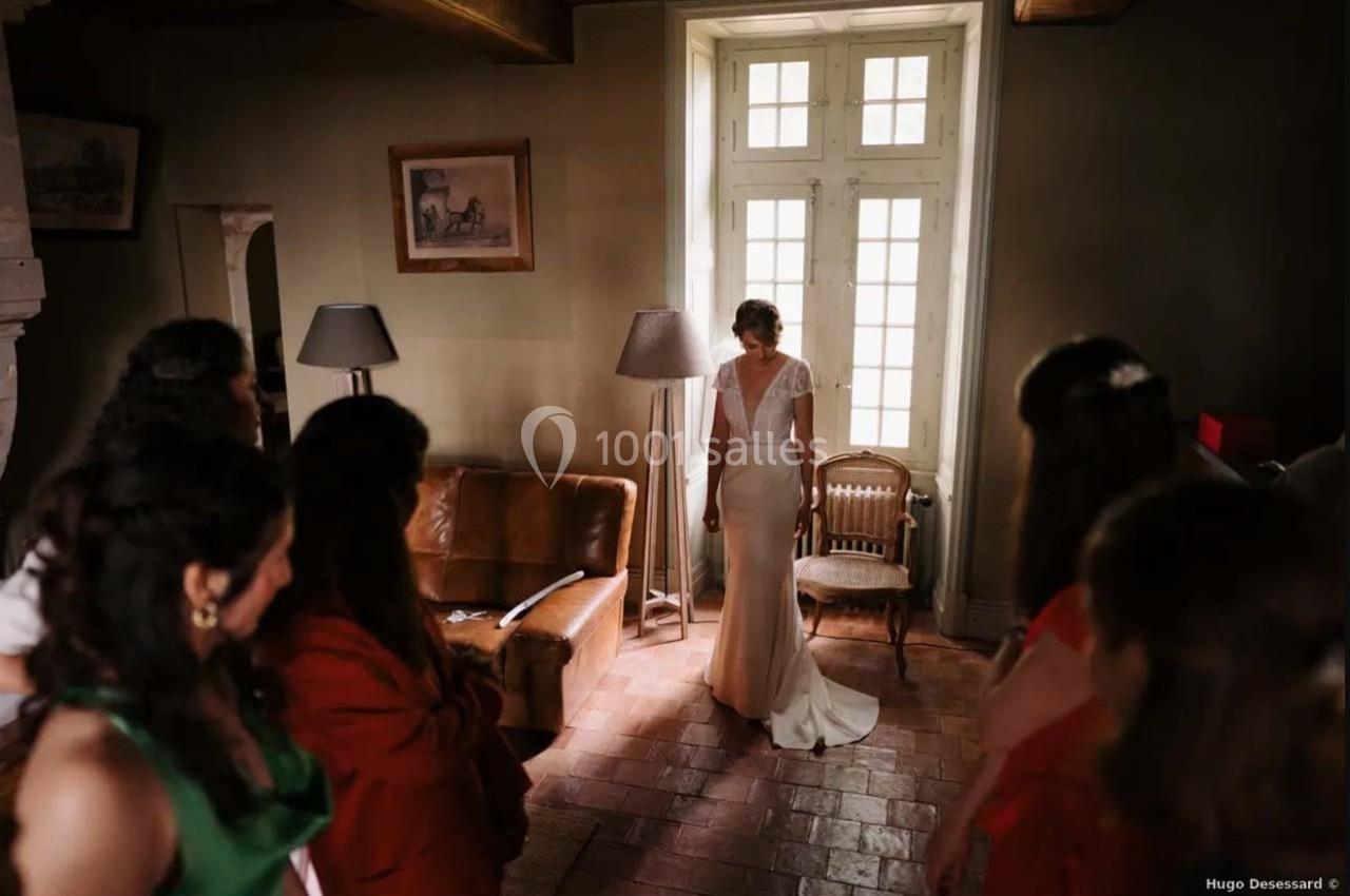 Une femme en robe blanche se tient debout près d'une fenêtre, entourée de personnes dans une pièce lumineuse.