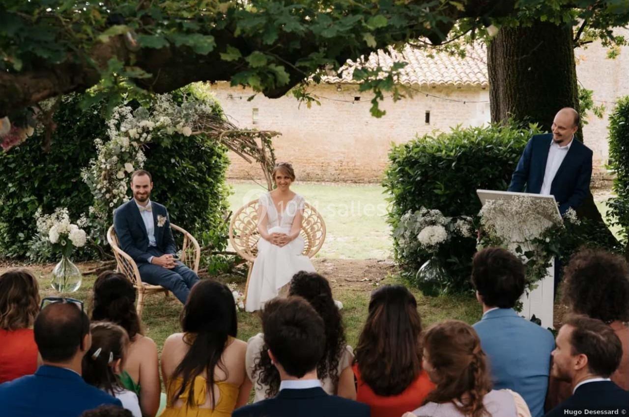 Un couple assis lors d'une cérémonie de mariage en extérieur, entouré d'invités et d'un officiant sous un arbre.