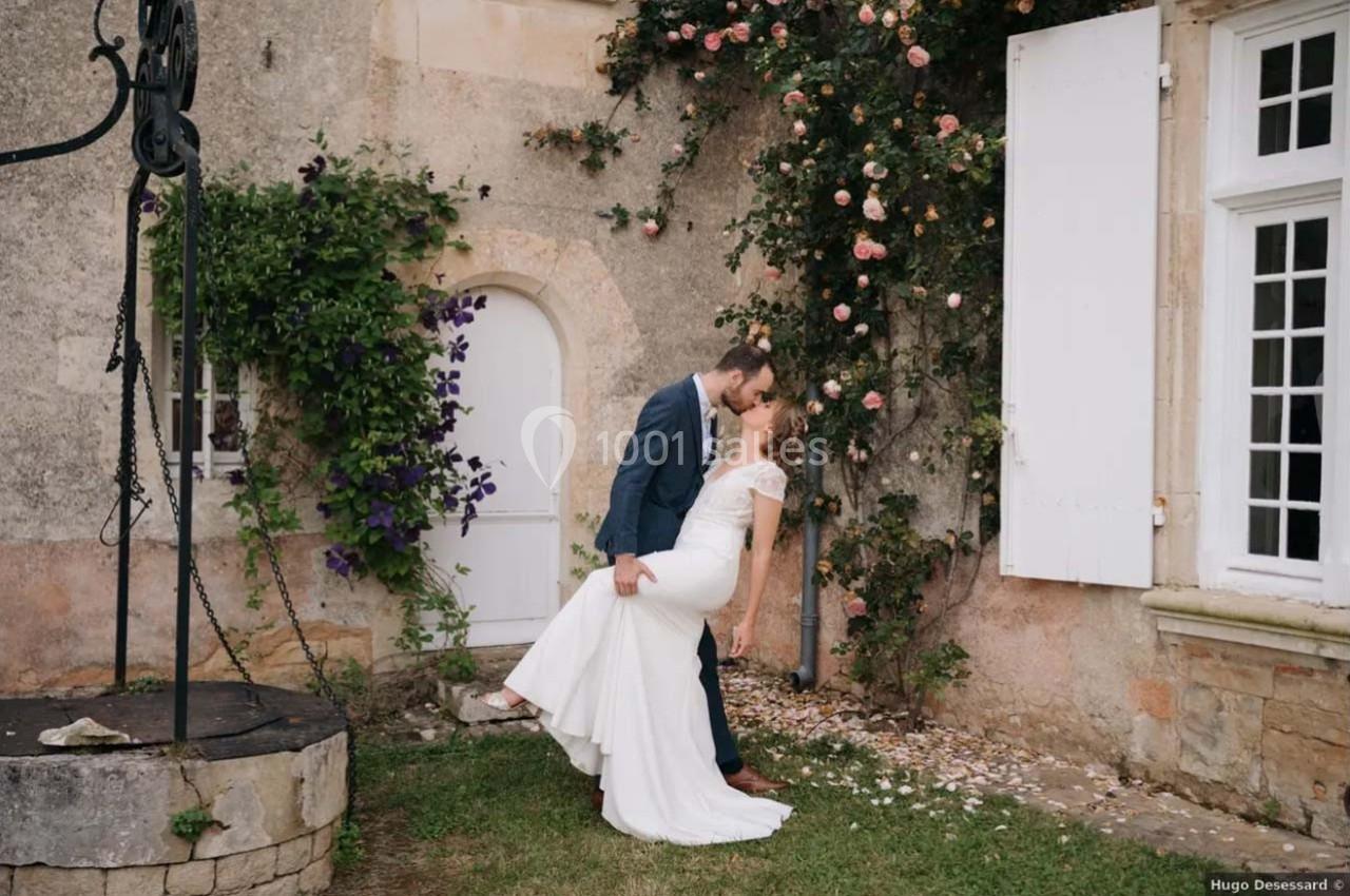 Un couple en tenue de mariage s'embrasse devant une façade en pierre ornée de rosiers et de volets blancs.