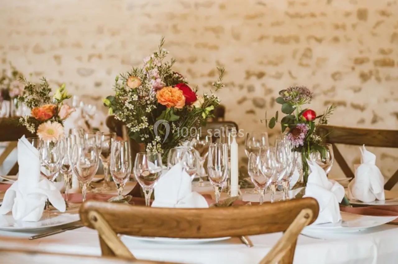 Table décorée avec des fleurs colorées, des verres en cristal et des serviettes pliées, dans une salle au mur en pierre.