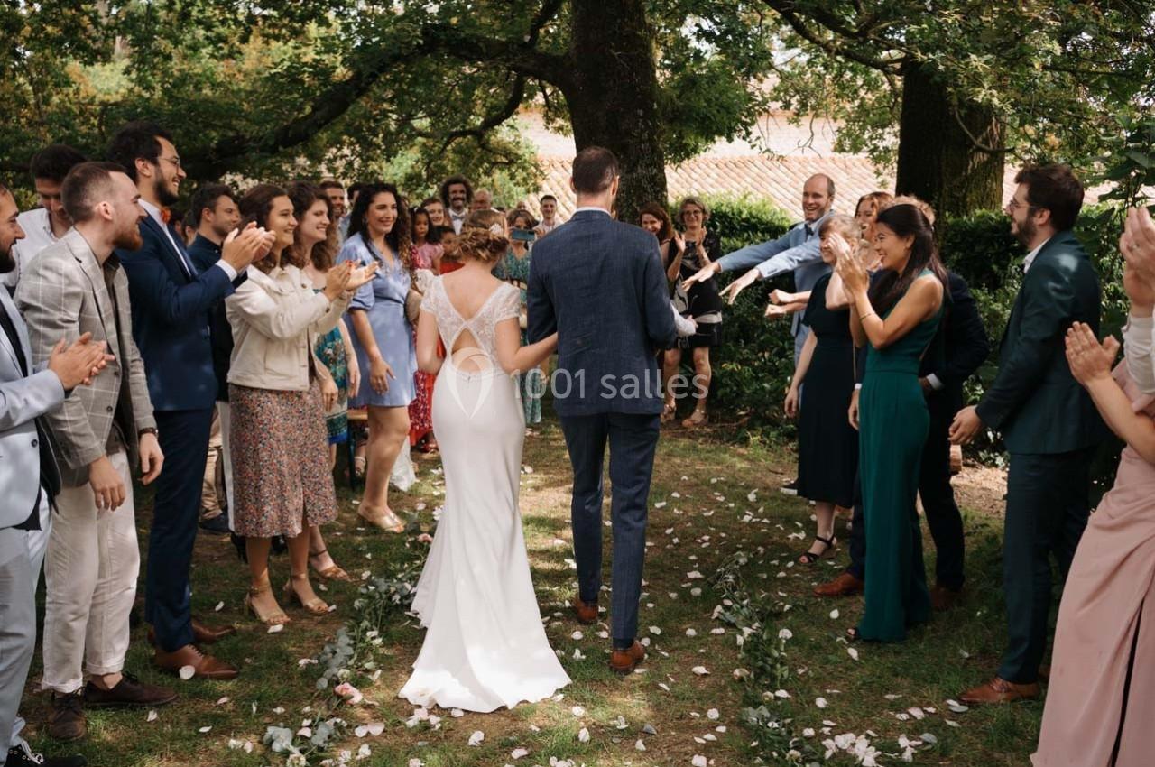 Un couple de mariés marche sur une allée de pétales, entouré d'invités applaudissant dans un jardin ombragé.