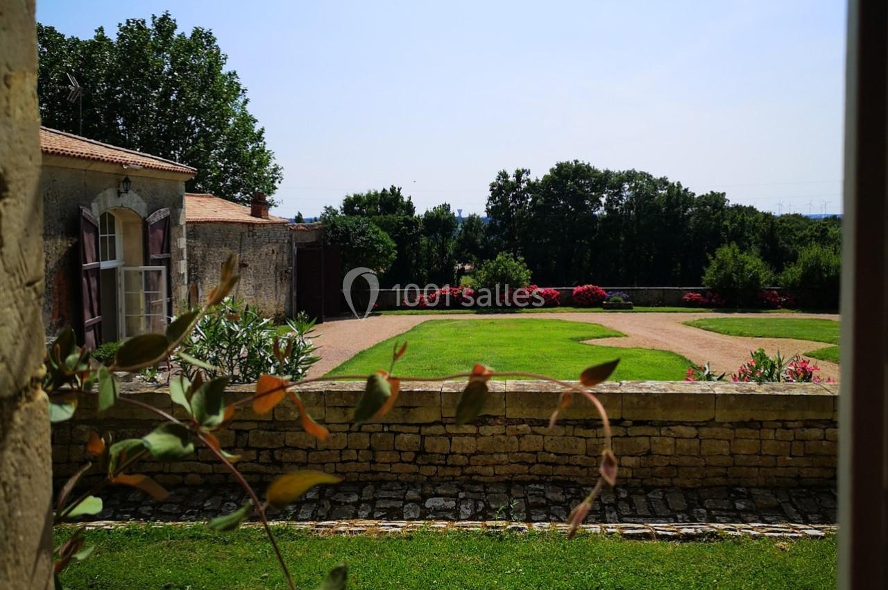 Vue sur un jardin avec pelouse, allées en gravier, parterre de fleurs rouges et bâtiments en pierre sous un ciel dégagé.