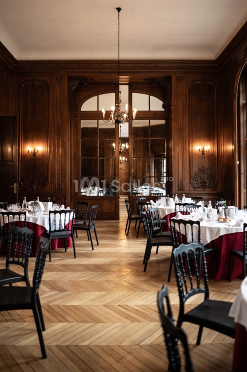 Salle élégante avec parquet en bois, murs lambrissés, tables dressées avec nappes blanches et chaises noires.