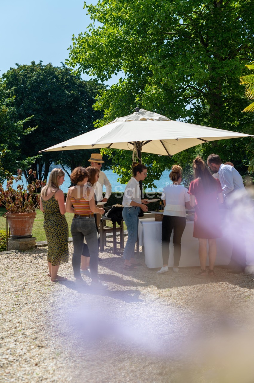 Groupe de personnes discutant près d'un stand sous un parasol dans un jardin ensoleillé.