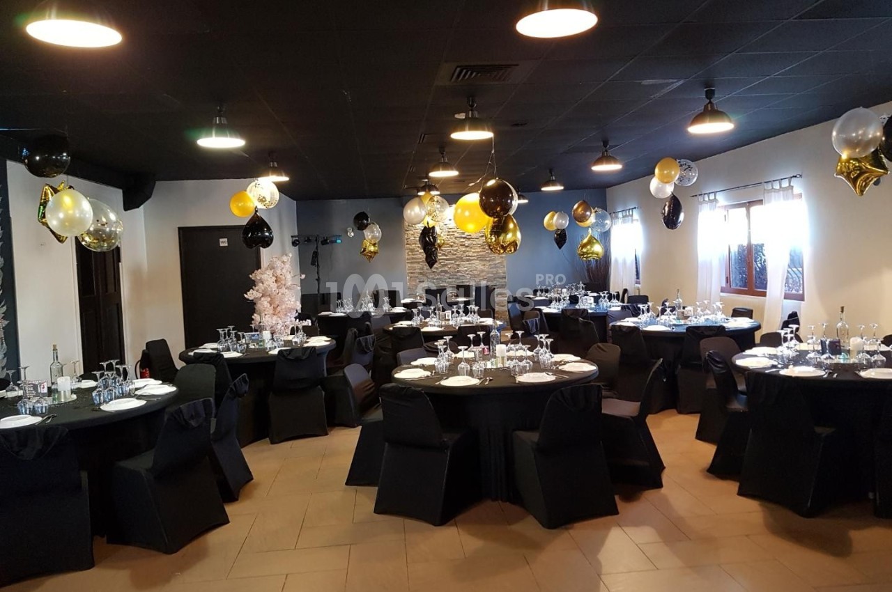 Salle décorée pour un événement avec des tables rondes dressées, des nappes noires et des ballons dorés, noirs et blancs.