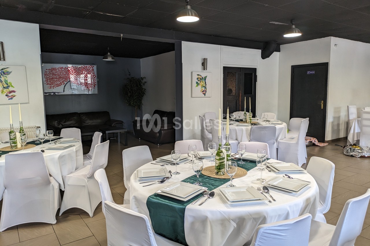 Salle de réception avec tables rondes dressées, nappes blanches, décorations vertes et chaises recouvertes.