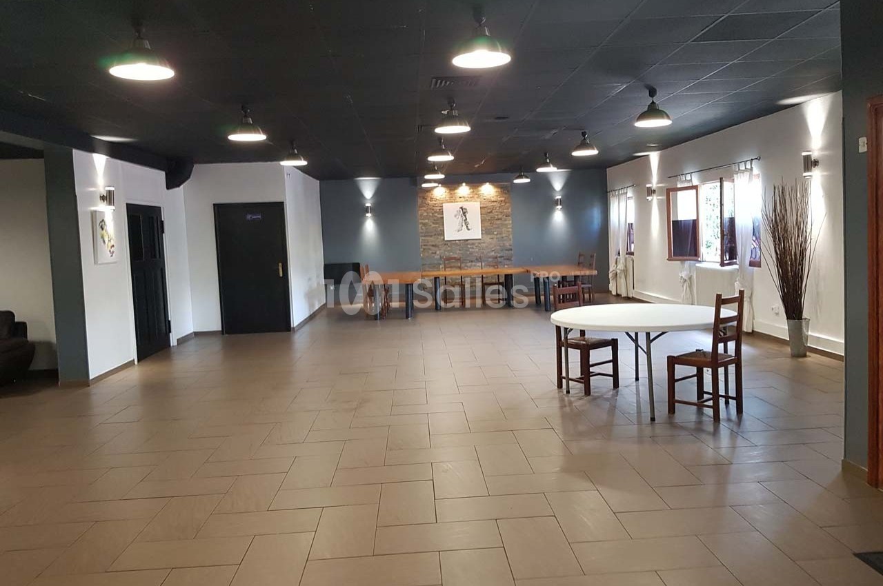 Salle spacieuse avec sol carrelé, plafond noir, tables et chaises dispersées, murs clairs et éclairage suspendu.
