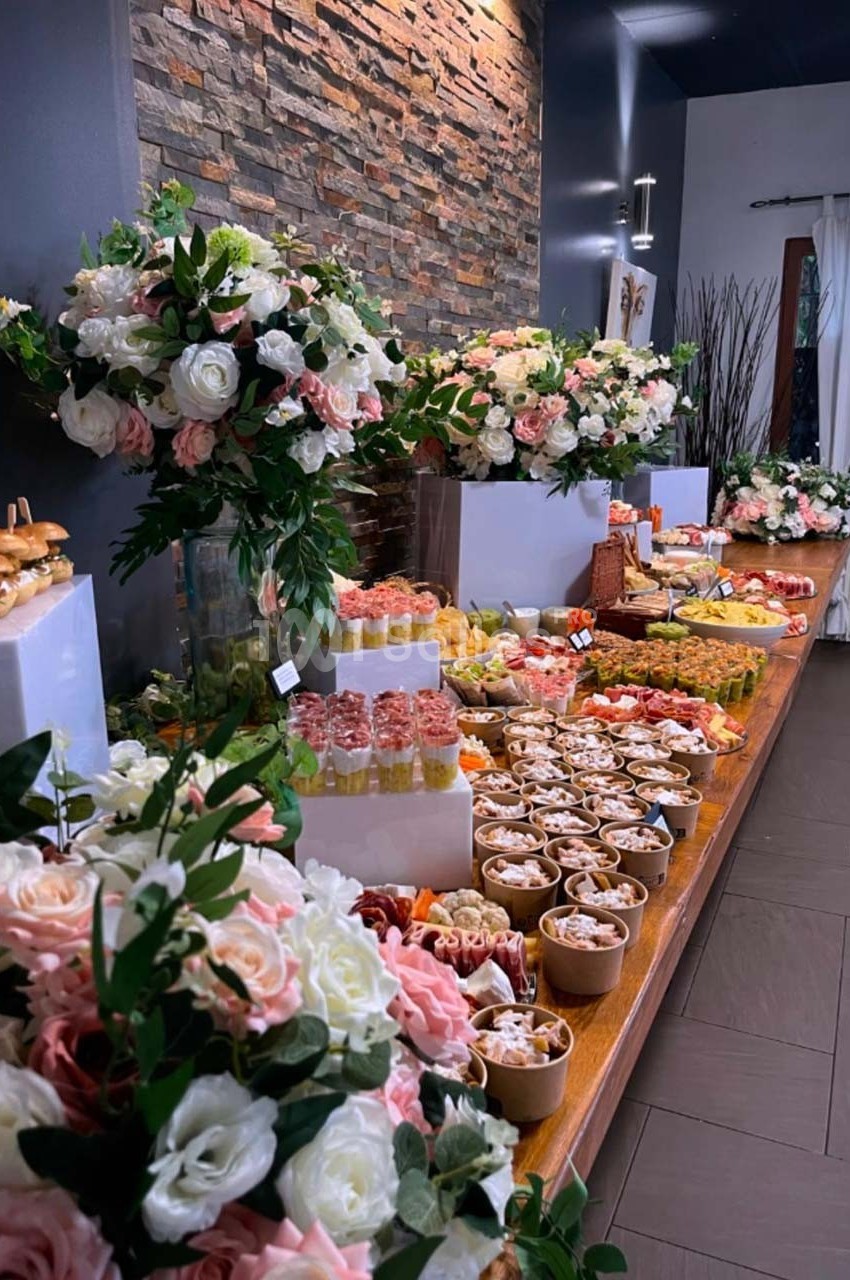 Table garnie de plats variés, desserts et fleurs décoratives, disposée dans une salle au mur de pierre.