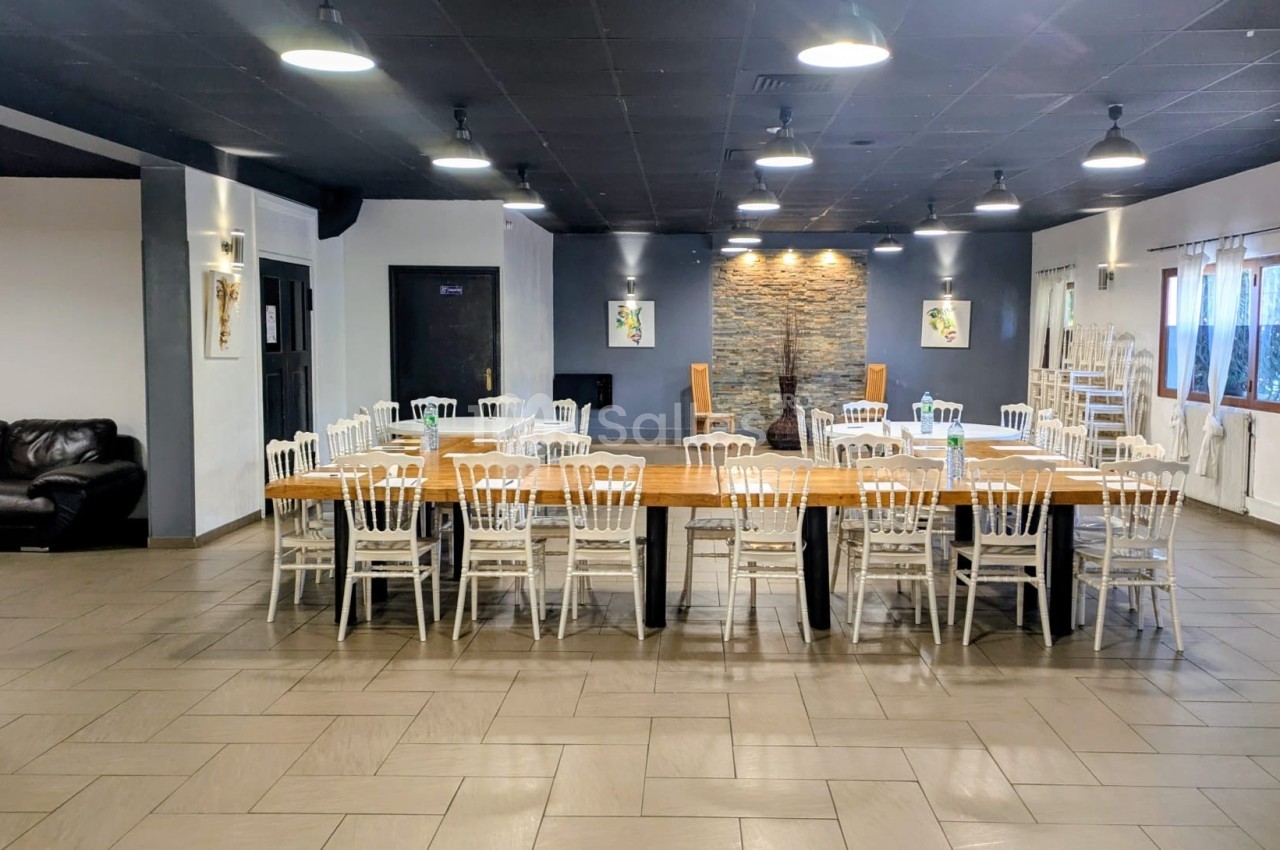 Salle spacieuse avec tables en bois alignées, chaises blanches, murs clairs et éclairage suspendu.