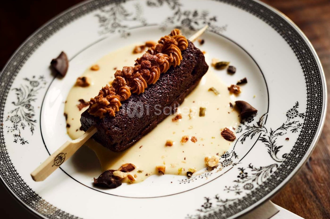 Dessert sur bâtonnet avec ganache au chocolat, noix concassées et sauce vanille, présenté sur une assiette décorée.