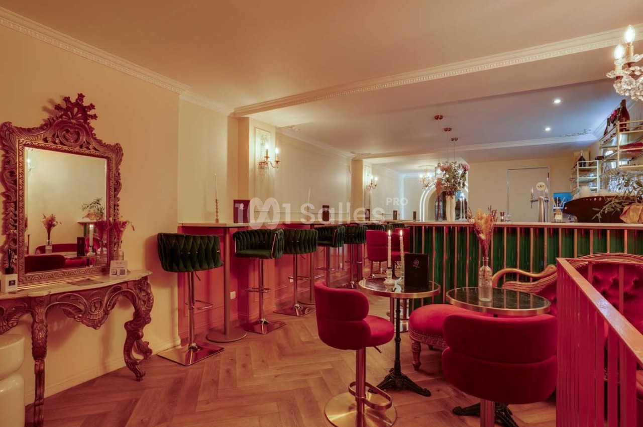 Intérieur d'un bar élégant avec tables, chaises en velours rouge, comptoir vert et miroir décoratif sur un mur beige.