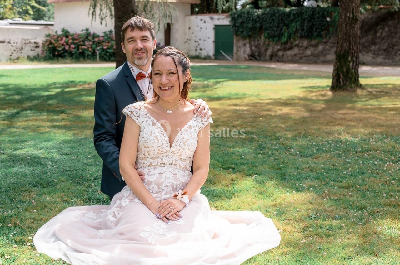 Un couple assis sur une pelouse dans un jardin, souriant devant un bâtiment clair entouré d'arbres.