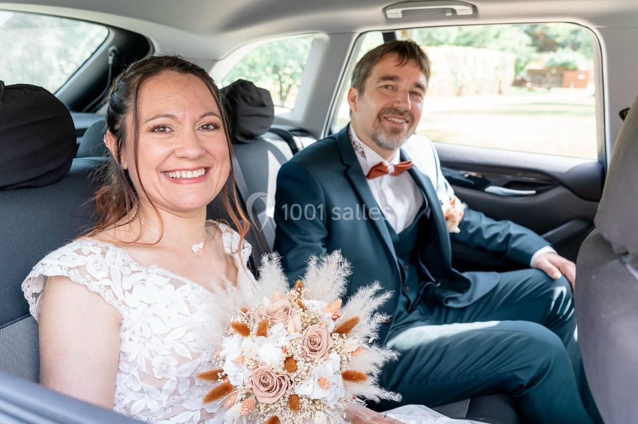 Un couple en tenue de mariage assis à l'arrière d'une voiture, la mariée tenant un bouquet de fleurs séchées.