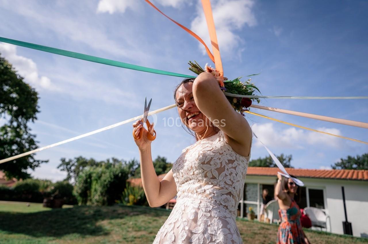 Une femme en robe blanche coupe des rubans colorés lors d'une célébration en extérieur par temps ensoleillé.