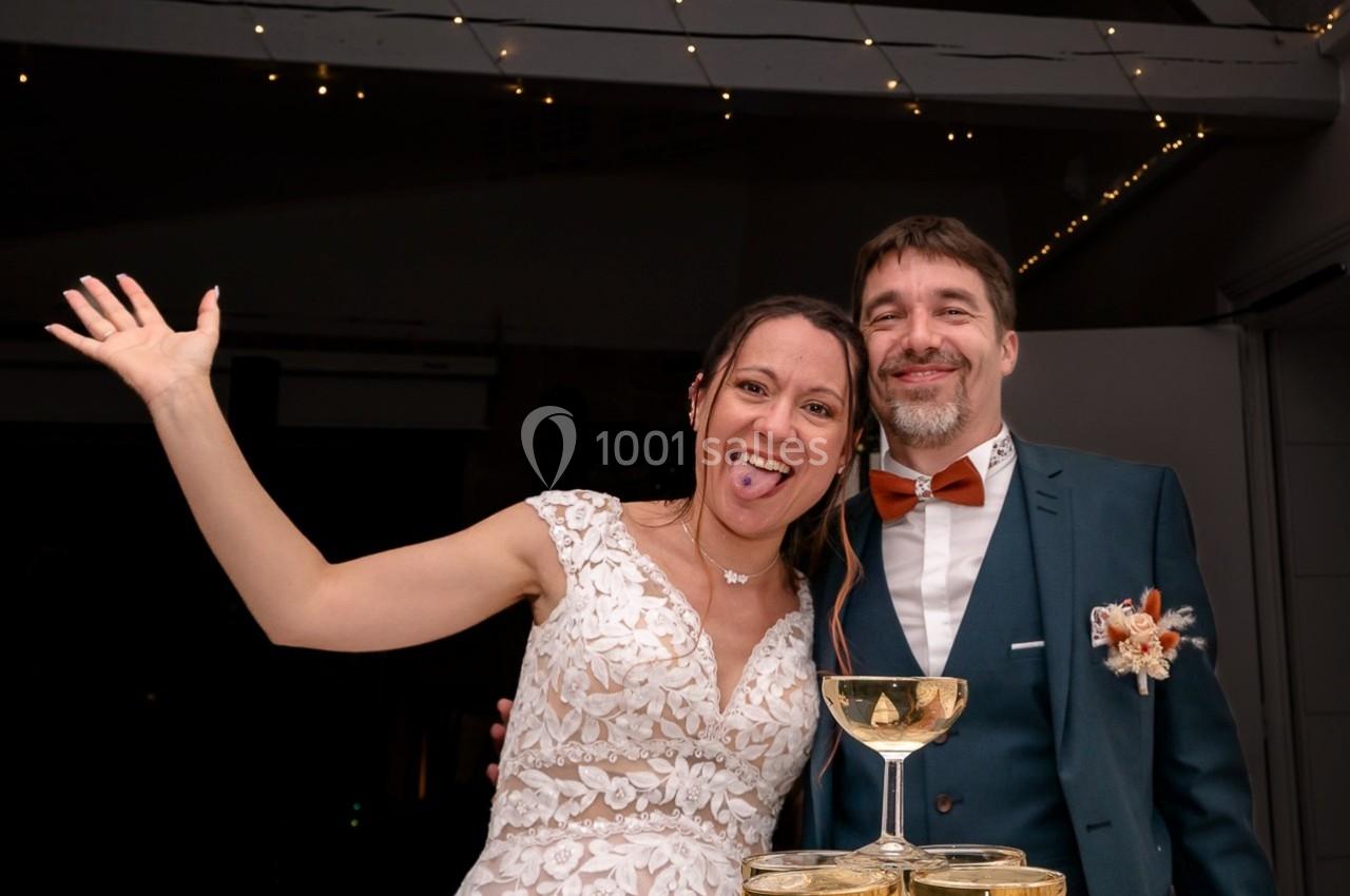 Un couple souriant en tenue de mariage pose devant une pyramide de coupes de champagne, sous des lumières festives.