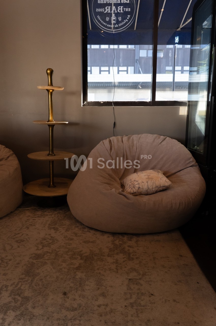 Fauteuil beige en forme de coussin avec un coussin décoratif, près d'une étagère en bois et d'une fenêtre.