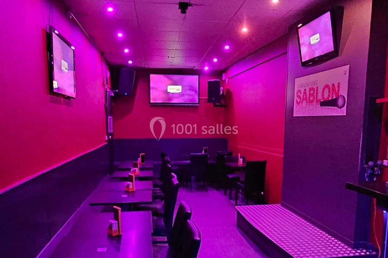 Salle de restaurant avec éclairage violet, tables alignées, écrans muraux et enseigne ’Sablon’ visible.