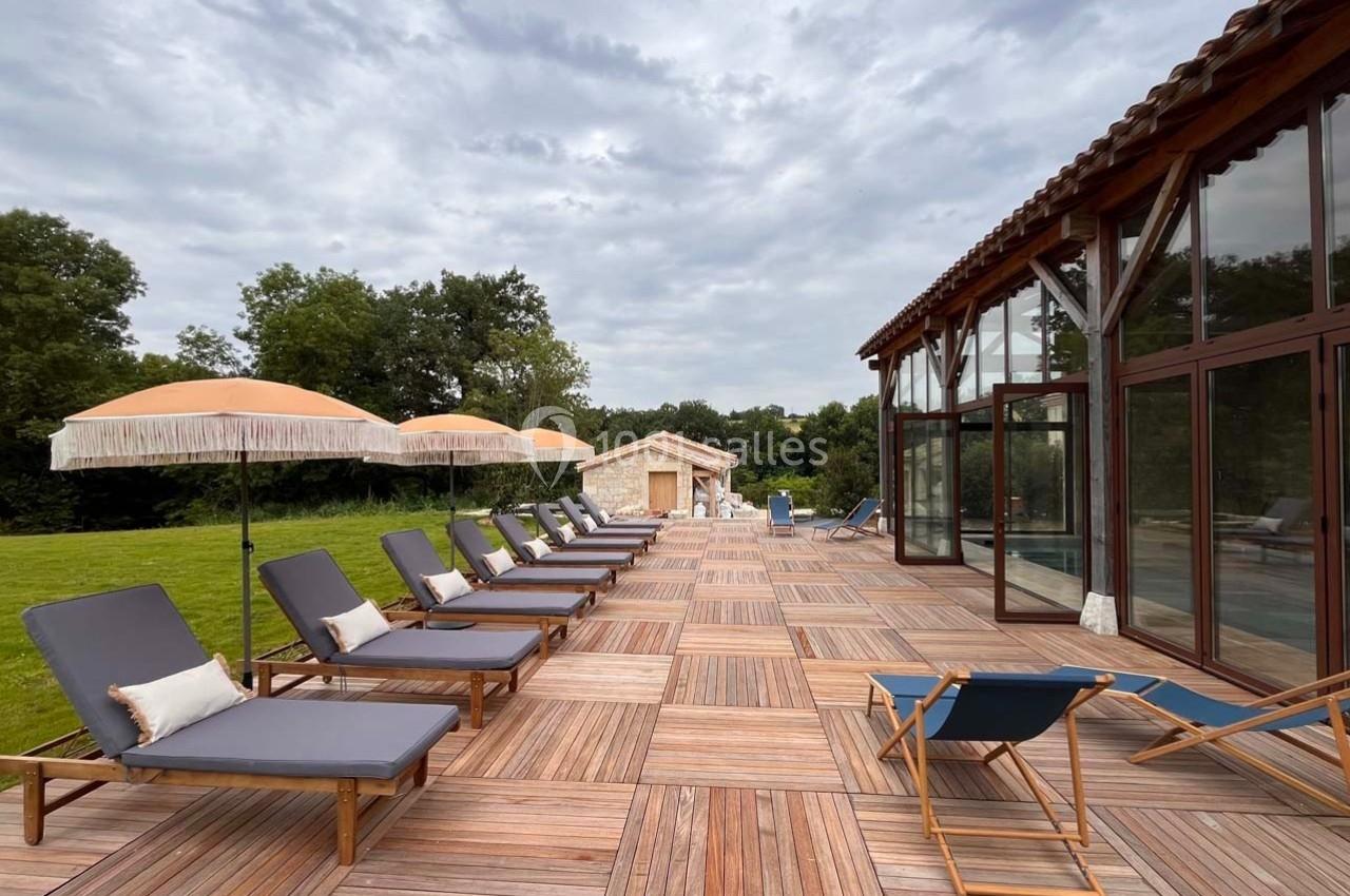 Terrasse en bois avec transats et parasols, bordée par une pelouse et un bâtiment vitré sous un ciel nuageux.