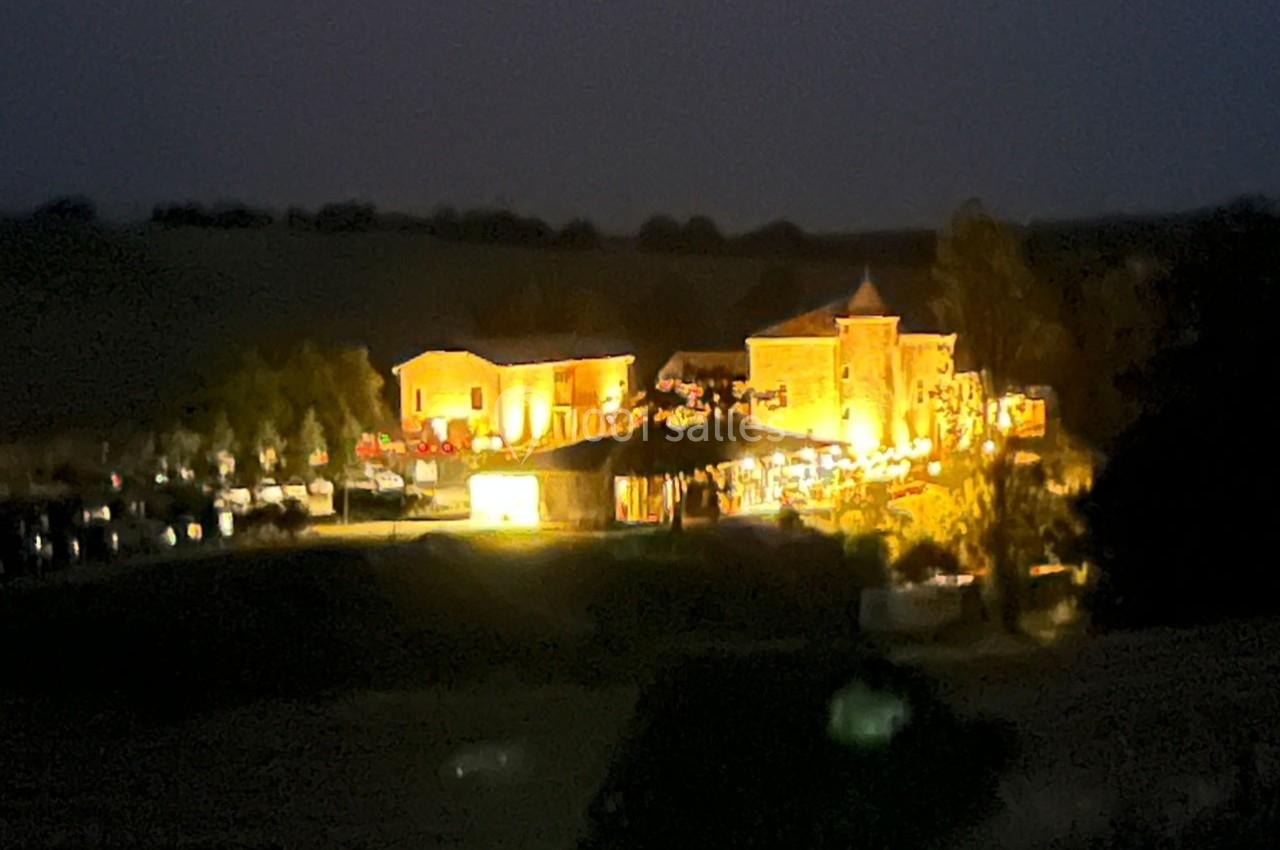 Bâtiments éclairés de nuit dans un paysage rural sombre, entourés de végétation et de quelques structures.
