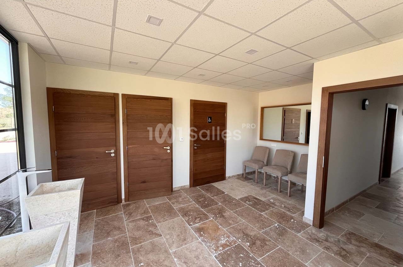 Salle avec sol en pierre, trois portes en bois, un miroir, et des chaises alignées contre un mur clair.