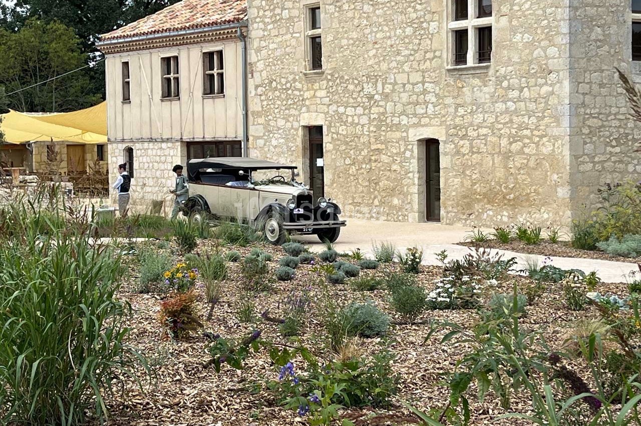Voiture ancienne stationnée devant un bâtiment en pierre, entouré d'un jardin avec des plantes et des fleurs.