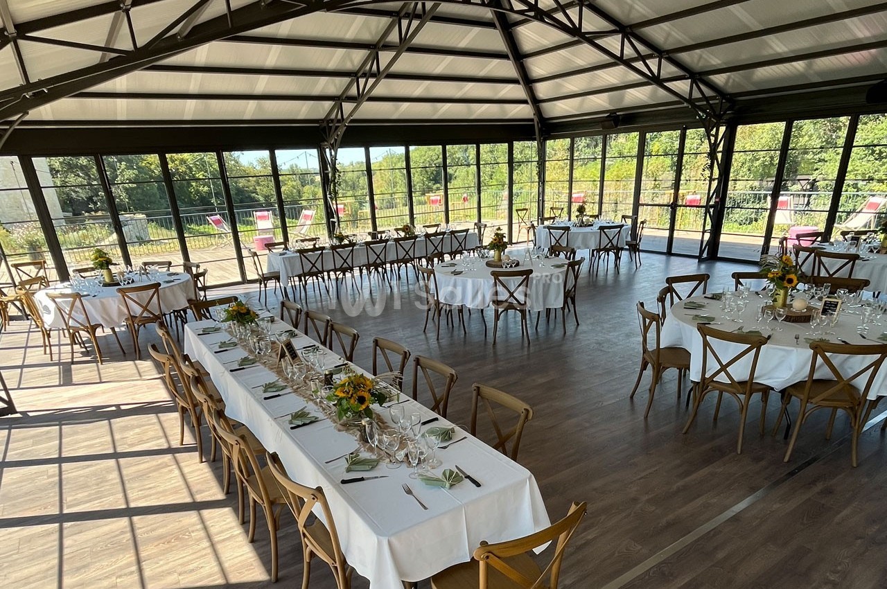 Salle lumineuse avec de grandes baies vitrées, tables dressées pour un repas, décorations florales sur les tables.