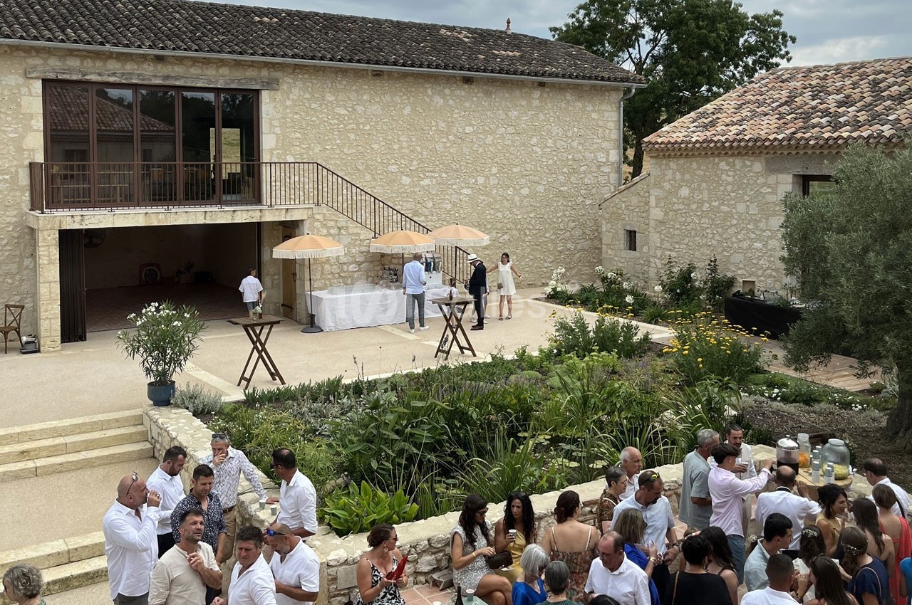 Groupe de personnes rassemblées dans la cour d'une bâtisse en pierre, avec un jardin et des parasols en arrière-plan.