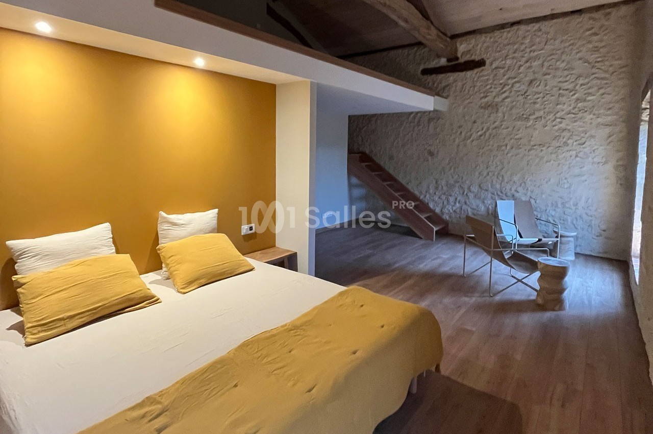 Chambre lumineuse avec un lit double, mur jaune, parquet en bois, escalier et coin détente avec fauteuils pliants.