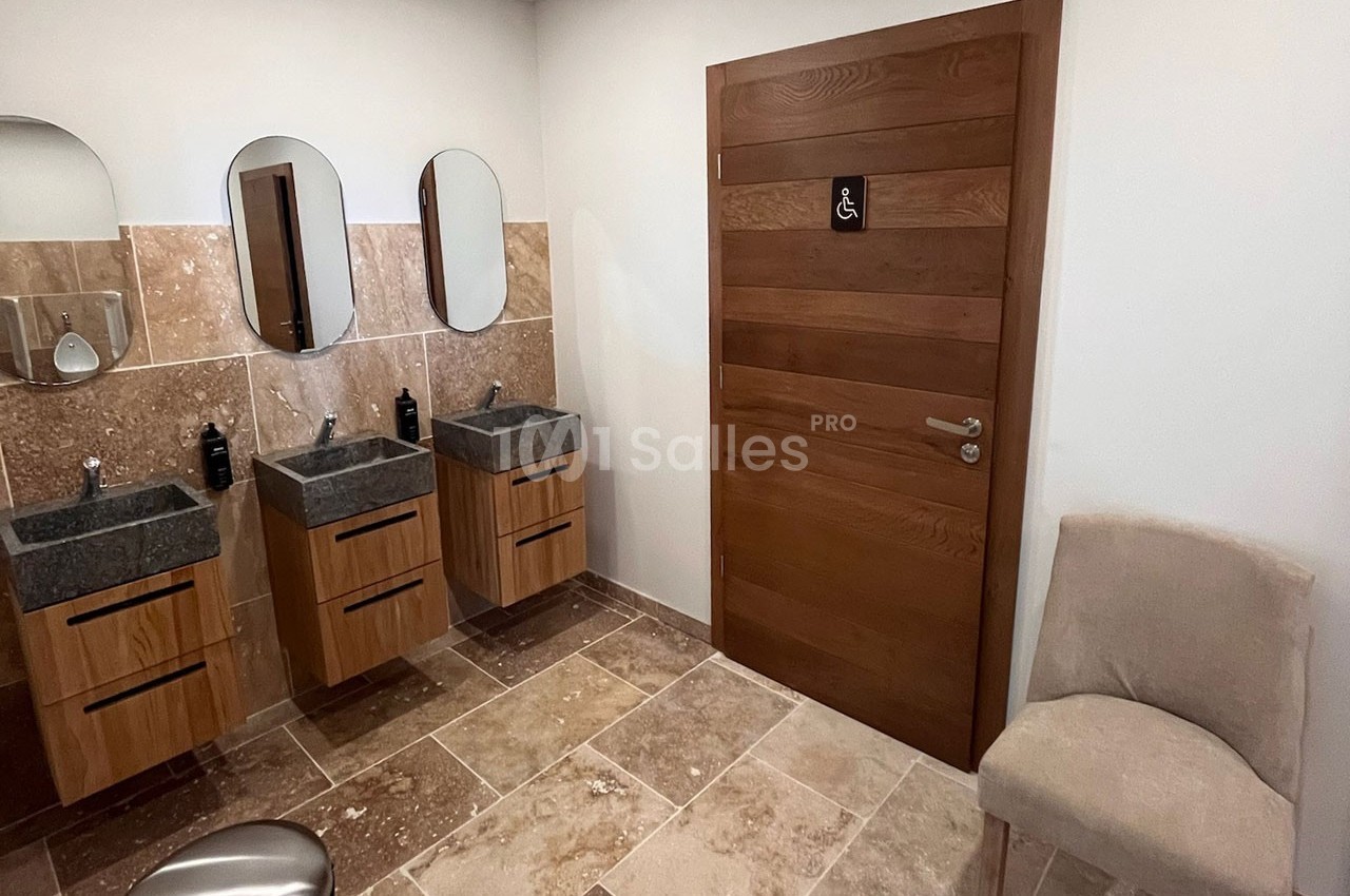 Salle de bain avec trois lavabos, miroirs ovales, sol en pierre et porte en bois avec pictogramme d'accessibilité.