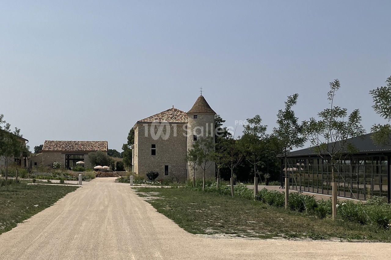 Chemin en gravier menant à un bâtiment en pierre avec une tour, entouré d'arbres et d'espaces verts.