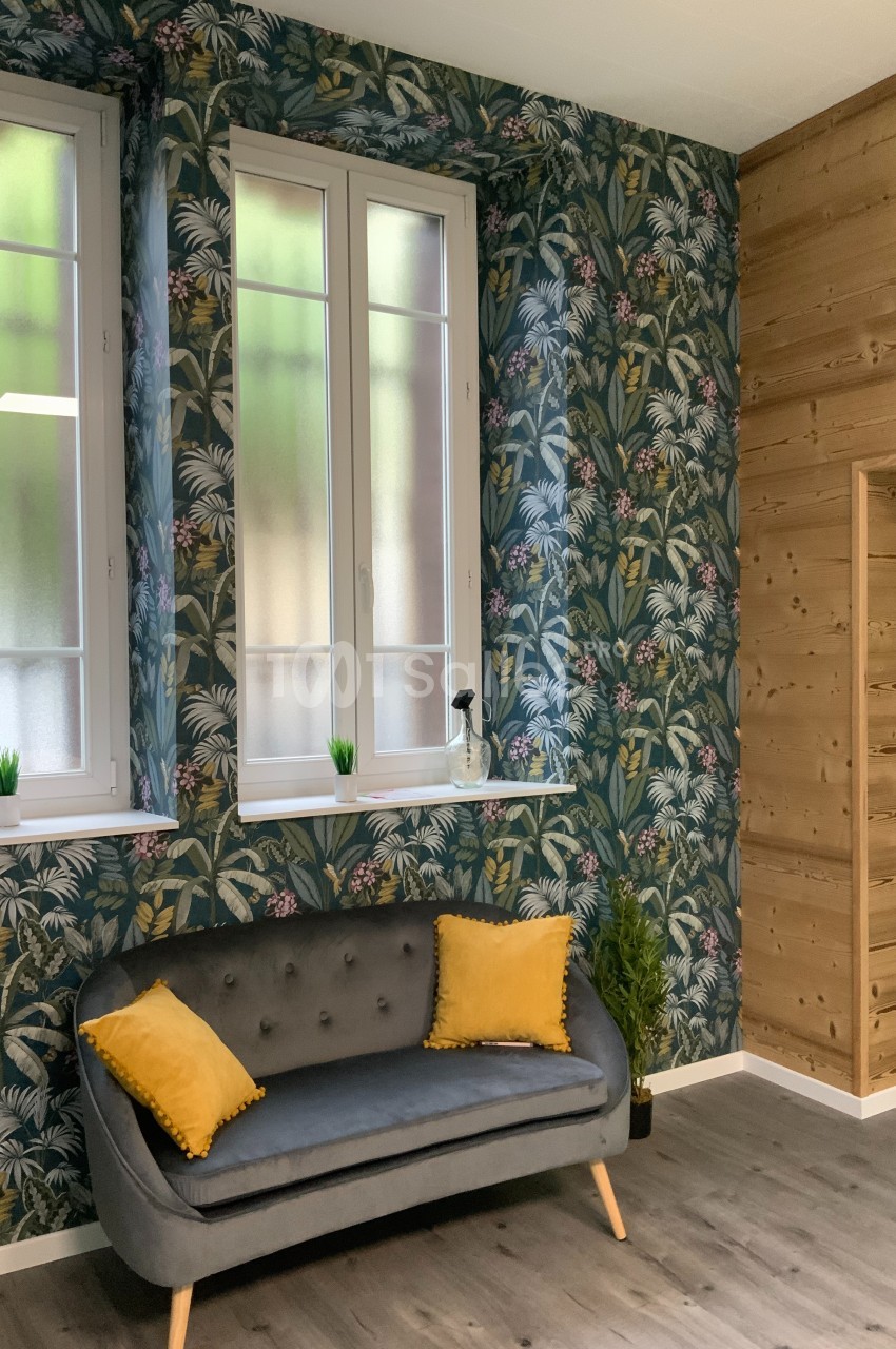 Salon avec canapé gris, coussins jaunes, mur tapissé de motifs tropicaux, lambris en bois et fenêtres vitrées.